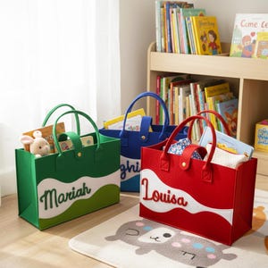 May include: Three felt tote bags in green, blue, and red, each with a white wave design and a name. The green bag says "Mariah", the blue bag says "Bright", and the red bag says "Louisa". Each bag has handles and contains books and toys.