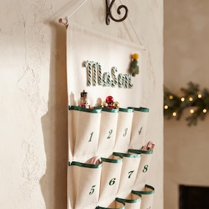 May include: A cream-coloured fabric advent calendar hanging from a metal hook. It features three rows of pockets with green trim, numbered one through twelve in green font. The name Mason is spelled out in candy cane-style lettering at the top.