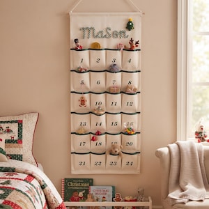 Op de afbeelding: Een crèmekleurige stoffen adventskalender voor aan de muur met vierentwintig vakjes, genummerd van één tot vierentwintig in zwarte letters. De naam Mason is bovenaan geborduurd in een zuurstokpatroon en de vakjes bevatten kleine kerstspeeltjes.
