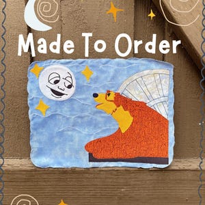 Peut inclure: Un article en tissu bleu rectangulaire avec un ours de dessin animé, une lune et des étoiles. L'ours est orange et jaune avec une aile. La lune a un visage souriant. Le texte "Made To Order" est au-dessus du dessin.