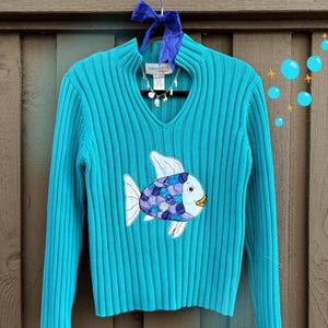 Könnte beinhalten: Türkisfarbener Rippstrickpullover mit V-Ausschnitt und langen Ärmeln. Der Pullover hat ein appliziertes Fischdesign mit blauen, lila und weißen Details. Eine silberne Halskette hängt am Ausschnitt. Der Pullover hängt an einem schwarzen Kleiderbügel mit blauem Band.
