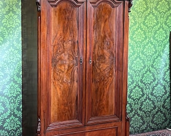 May include: A tall, antique wooden wardrobe with two doors and a drawer. The wardrobe is made of dark brown wood with a natural wood grain pattern. The doors have ornate carvings and brass hardware. The wardrobe is a classic piece of furniture.