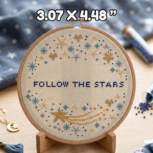 Peut inclure: Une broderie au point de croix dans un cercle en bois, mesurant 7,8 x 11,4 cm. Le motif présente un thème céleste avec des étoiles et le texte "FOLLOW THE STARS" en bleu marine. Les étoiles sont dans des tons de jaune et de bleu.