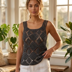 May include: A dark gray crochet tank top with a square neckline and diamond-shaped openwork design. The top has thin straps and a slightly rounded hem. The model is wearing white pants. The background includes plants and natural light.