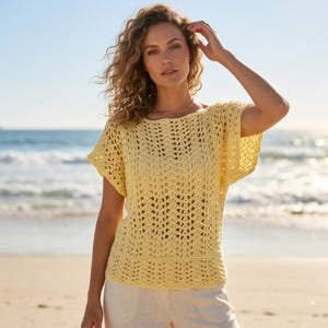 May include: A light yellow crochet top with short sleeves. The top has an open, airy design and a round neckline. The model is wearing the top on a beach with the ocean in the background. The top is paired with white pants.