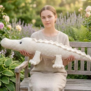 May include: A large, cream-colored crochet alligator plush toy with a detailed, textured design. The alligator has a long body, four small feet, and a row of decorative scales along its back. The toy is being held by a person sitting on a wooden bench.
