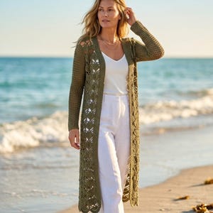 May include: A long, olive green crochet cardigan with openwork detailing. The cardigan is worn over a white top and white pants. The photo is taken on a beach with the ocean in the background.