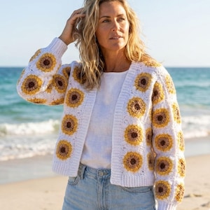 May include: A white crochet cardigan featuring a sunflower pattern in yellow and brown. The open-front sweater has long sleeves and is worn over a white top and blue jeans. The cardigan is a handmade item.