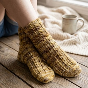 May include: A pair of ribbed knit socks in shades of brown and yellow. The socks are thick and appear warm, with a textured pattern. A neutral-coloured mug and a cream-coloured blanket are in the background, suggesting a cosy setting.