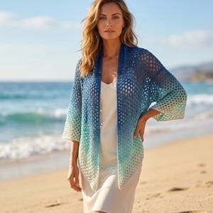 May include: A woman wearing a blue, open-weave cardigan with a gradient effect, transitioning from dark blue to light blue and white. The cardigan has three-quarter sleeves and is worn over a white dress. The background shows a beach and ocean.