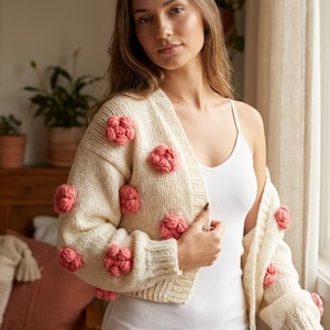May include: Cream-colored, open-front cardigan sweater adorned with multiple coral-colored, three-dimensional flower appliques. The sweater is made of a chunky knit material and has long sleeves. The model is wearing a white tank top underneath.
