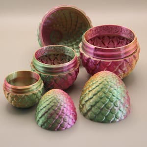 May include: A selection of iridescent dragon egg-shaped containers in different sizes. The containers are mainly pink, green, and gold, with a textured, scale-like design. The set includes two halves of a large egg, two lidded containers, and two smaller egg-shaped pieces.