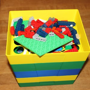 Könnte beinhalten: Ein gelber, grüner und blauer Aufbewahrungsbehälter gefüllt mit bunten Spielzeugbausteinen. Die Bausteine sind rot, blau, grau und grün. Eine grüne Grundplatte befindet sich im Inneren des Behälters.