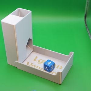 Könnte beinhalten: Ein weißer Würfelturm mit einem Tablett und einem blauen Glitzerwürfel mit weißen Zahlen. Das Tablett hat goldenen Text mit der Aufschrift "Lygon Vendor". Der Turm steht auf einer grünen Oberfläche.