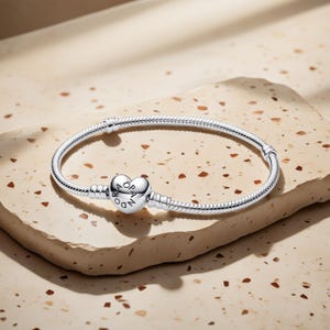 Puede incluir: Una pulsera plateada con un cierre en forma de corazón. La pulsera tiene un diseño de cadena de serpiente y se muestra sobre una superficie beige. El cierre de corazón tiene grabado el nombre de la marca. Joyería.