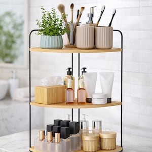 May include: A three-tiered bathroom shelf with a black metal frame and light wood shelves. The top shelf holds a potted plant, brushes, and beige ceramic cups. The middle shelf has skincare products, and the bottom shelf has cosmetics.