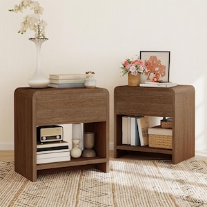 Puede incluir: Dos mesitas de noche de madera oscura a juego con bordes superiores redondeados, cada una con un cajón y un estante inferior abierto. La mesita izquierda tiene un jarrón blanco con flores, libros y una radio. La derecha tiene un cuadro, flores y libros.