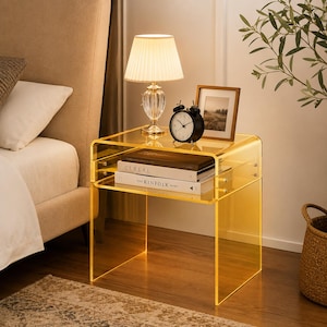 May include: A transparent yellow acrylic side table with a U-shaped design and a lower shelf. On the table sits a lamp with a pleated shade, a black analog alarm clock, and a picture frame. Two books titled Cereal and The Kinfolk Home are stacked.