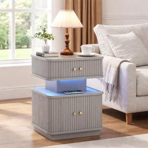 May include: A two-tiered gray nightstand with vertical grooved detailing and gold-colored knobs. The top tier holds a lamp, a book, and a small vase. The bottom tier has a blue LED light and a smartphone. The nightstand is next to a sofa.