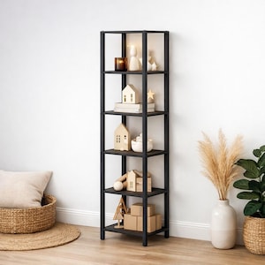 May include: A tall, black, open-frame bookshelf with five shelves. The shelves display decorative items including small wooden houses, candles, books, and a star. The shelf is against a white wall, with a woven basket and a plant in the foreground.