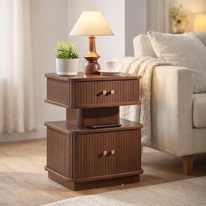 May include: A two-tiered, dark brown wooden side table with a lamp, a small potted plant, and a teacup. The table has two drawers with gold-colored knobs. The lamp has a beige lampshade. The table is next to a beige sofa.