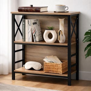 Puede incluir: Una estantería de tres niveles con acabado de vetas de madera clara y marco de metal negro. La balda superior contiene un libro, una taza de café y un posavasos. La balda central exhibe libros y jarrones decorativos. La parte inferior tiene una cesta, una máscara para los ojos y servilletas.