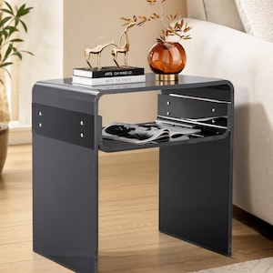 May include: A modern, dark gray side table with a clear glass top and a lower shelf. The table features a curved design and holds decorative items, including a gold deer figurine, books, and a glass vase with a plant. The table is placed next to a beige sofa.