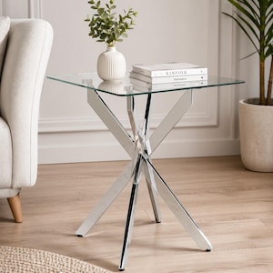 May include: A modern side table with a square glass top and a polished chrome base. The base features an X-shaped design. A white vase with greenery and two books are on the table.