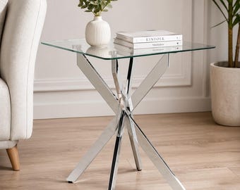 May include: A modern side table with a square glass top and a polished chrome base. The base features an X-shaped design. A white vase with greenery and two books are on the table.