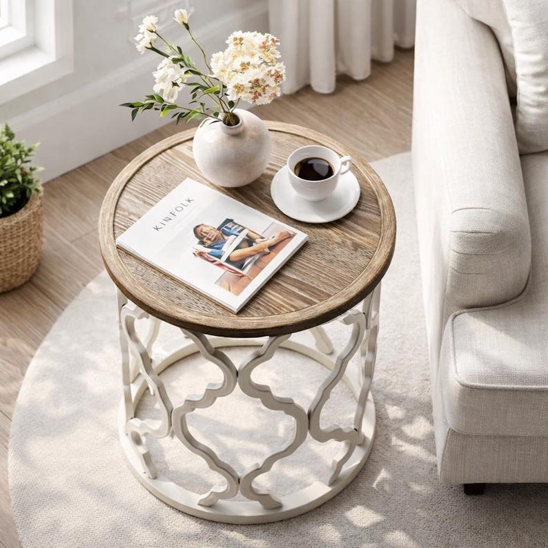 Puede incluir: Una mesa auxiliar redonda de madera con base de metal blanco. La mesa tiene una tapa de color marr&oacute;n claro con un borde decorativo. Un jarr&oacute;n blanco con flores, una taza de caf&eacute; y una revista est&aacute;n sobre la mesa. La revista se titula "KINFOLK".