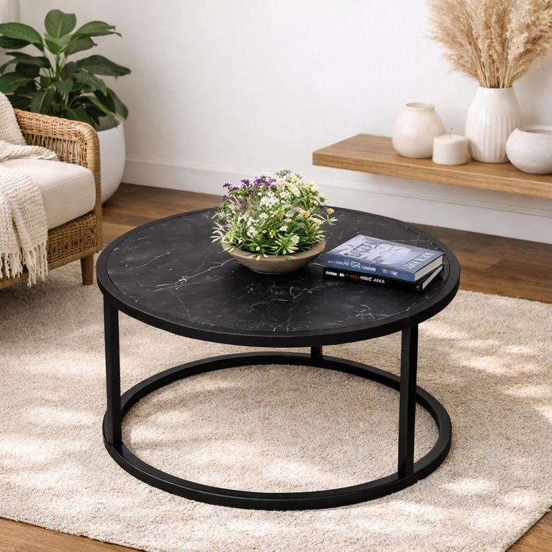 May include: A round coffee table with a black marble-look top and a black metal frame. The table has a circular base and straight legs. A small bowl of flowers and a book are on the table. The table is on a light-colored rug.
