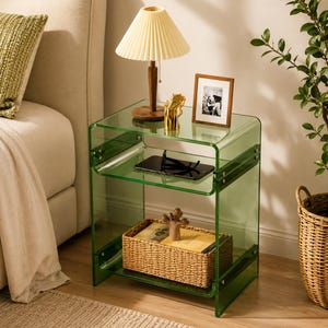 May include: A transparent green acrylic side table with two shelves, featuring metal hardware connectors. The top shelf holds a pleated lamp, a gold figurine, and a framed photo. The middle shelf holds a black tablet and glasses, while the bottom shelf contains a woven basket.