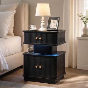 May include: A black nightstand with two drawers, each with gold-colored knobs. A white lamp, a framed picture, and a coffee cup sit on top. A tablet is charging on the middle shelf.