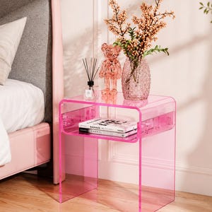 May include: A translucent pink acrylic side table with a curved top and a lower shelf. On the table sits a clear glass vase with peach-colored berries, a pink bear figurine, a reed diffuser, and two books titled Bauhaus and Chanel.