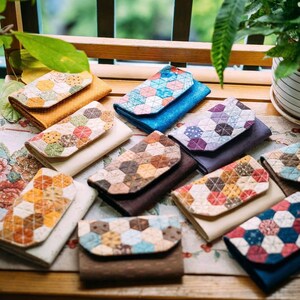 May include: A collection of handmade wallets with colorful, geometric patchwork designs. The wallets feature a variety of colors, including blue, brown, orange, and cream. The patchwork patterns are made of small, individual fabric pieces.