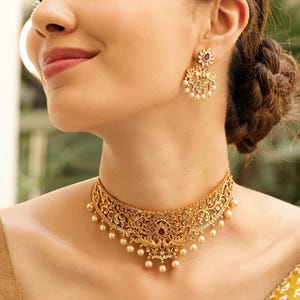 Op de afbeelding: Een goudkleurige ketting en bijpassende oorbellen set. De ketting is een choker-stijl met ingewikkelde filigraan en parel accenten. De oorbellen hebben een bloemenontwerp met een rode edelsteen in het midden en bungelende parels. De sieraden worden gedragen.