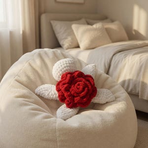 May include: A cream-colored beanbag chair with a large, handmade red and white crochet flower. The flower has a detailed, textured design, with the red petals in the center and white petals surrounding it. The beanbag is in a bedroom setting.