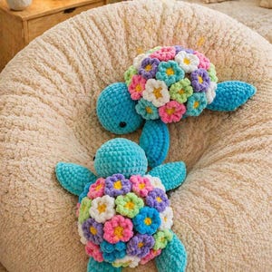 Könnte beinhalten: Zwei handgefertigte, türkisfarbene Häkelschildkröten-Plüschtiere. Die Schildkrötenpanzer sind mit bunten Häkelblumen in Rosa, Weiß, Gelb, Lila und Grün verziert. Die Schildkröten liegen auf einer beigefarbenen, strukturierten Oberfläche.