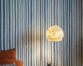 May include: A wall-mounted pendant light with a brass-colored metal arm and a textured, translucent, off-white spherical shade. The light hangs vertically, emitting a warm glow, and features a cord that runs down the wall to a power outlet.