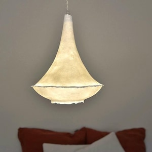 May include: A cream-coloured pendant light with a bell-shaped shade. The shade has a textured surface and a wide, slightly ruffled bottom edge. The light hangs from a white cord, illuminating a bedroom setting.