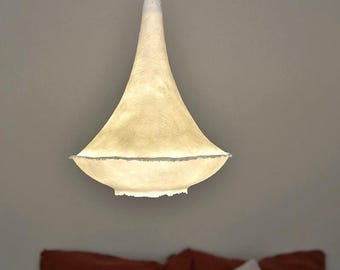 May include: A pendant light fixture with a cream-colored, bell-shaped shade. The shade has a textured surface and a wide, slightly ruffled bottom edge. The light hangs from a white cord, illuminating a bedroom setting.