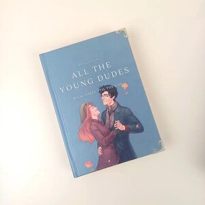 Puede incluir: Un libro de tapa dura titulado "ALL THE YOUNG DUDES" con el subt&iacute;tulo "BOOK THREE TIL THE END" en una cubierta azul claro. La portada presenta una pareja bailando, con hojas de oto&ntilde;o. El libro tiene detalles dorados en las esquinas.