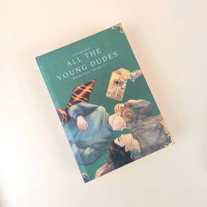 Puede incluir: Un libro de tapa dura titulado "ALL THE YOUNG DUDES BOOK TWO: YEARS 5-7" con una cubierta verde azulado. La portada presenta una ilustraci&oacute;n de dos personas tumbadas, una bufanda y una linterna. El libro tiene dise&ntilde;os decorativos en las esquinas.