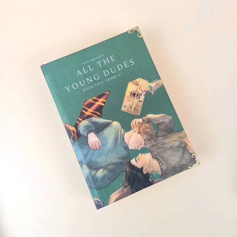 Puede incluir: Un libro de tapa dura titulado "ALL THE YOUNG DUDES BOOK TWO: YEARS 5-7" con una cubierta verde azulado. La portada presenta una ilustraci&oacute;n de dos personas tumbadas, una bufanda y una linterna. El libro tiene dise&ntilde;os decorativos en las esquinas.