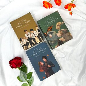 Puede incluir: Tres libros titulados "All The Young Dudes" se muestran sobre una superficie blanca. Los libros tienen cubiertas de diferentes colores: dorado, verde y azul. Cada libro presenta ilustraciones y texto. Tambi&eacute;n hay una rosa roja y flores naranjas.