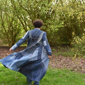 May include: A long, floor-length coat constructed from various shades of blue denim fabric in a patchwork design. The garment features a structured collar, long sleeves, and a lace-up detail on the upper back, with visible seams and pockets.