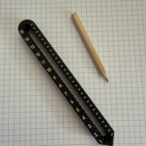 Könnte beinhalten: Ein schwarzes Lineal mit gelben Markierungen, nummeriert von 1 bis 15, und ein Bleistift aus Holz. Das Lineal ist schräg auf einem karierten Papier angeordnet. Der Bleistift ist gespitzt und liegt parallel zum Lineal.