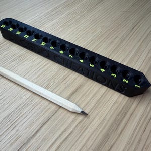 Puede incluir: Un portalápices negro con 15 agujeros numerados y el texto "GEEGO CREATIONS" en verde. Un lápiz blanco se coloca junto al soporte. El soporte está sobre una superficie de madera de color marrón claro.
