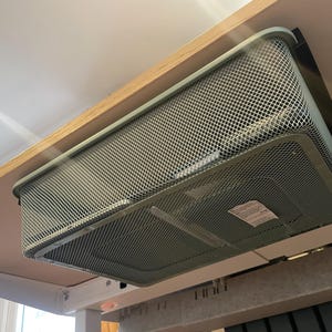 May include: A gray metal mesh storage basket mounted under a wooden desk. The basket is rectangular with rounded corners and a grid pattern. The desk is light-colored, and the basket is attached with a black bracket.