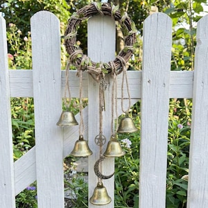 May include: A rustic wreath made of woven twigs and green leaves hangs on a white picket fence. Below, five brass bells are suspended by twine, along with a key, a star, and a crescent moon charm. The bells are of varying sizes.
